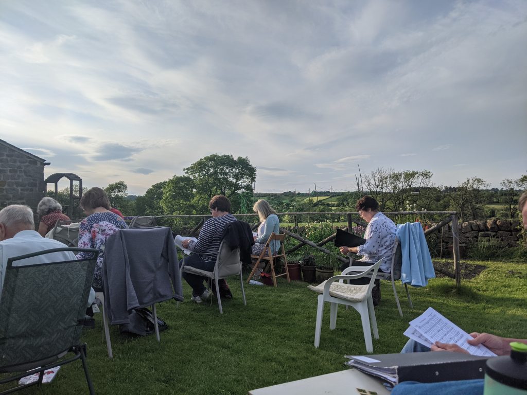 Some members of the choir at a socially distanced rehearsal in a garden, with a view over the countryside beyond.